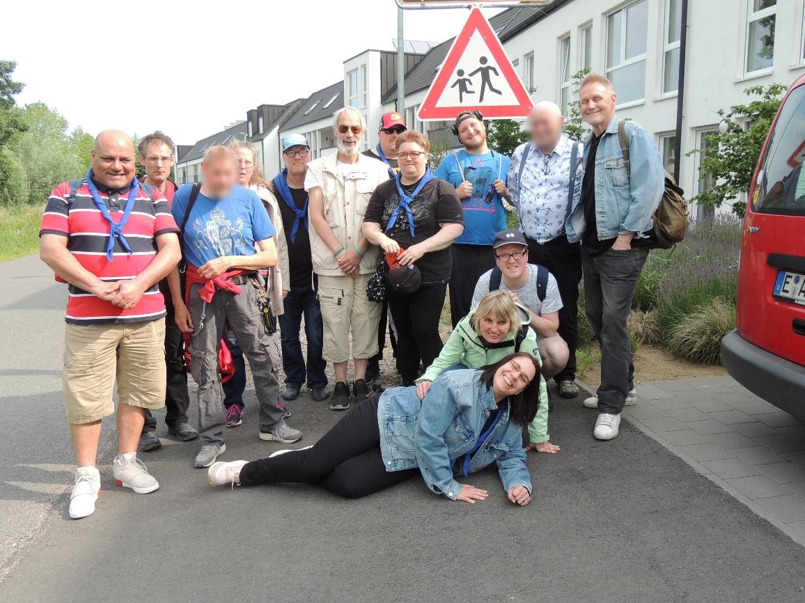 Gruppenbild von der Pfingstfahrt 2018 nach Bocholt-Suderwick: 14 Pfadfinderinnen und Pfadfinder in bunter Alltagskleidung. Einige tragen ein blaues Halstuch. 11 Personen stehen nebeneinander. Davor sind zwei Personen in der Hocke. Eine Person liegt quer vor den anderen. Im Hintergrund ist eine Wohnsiedlung zu sehen.