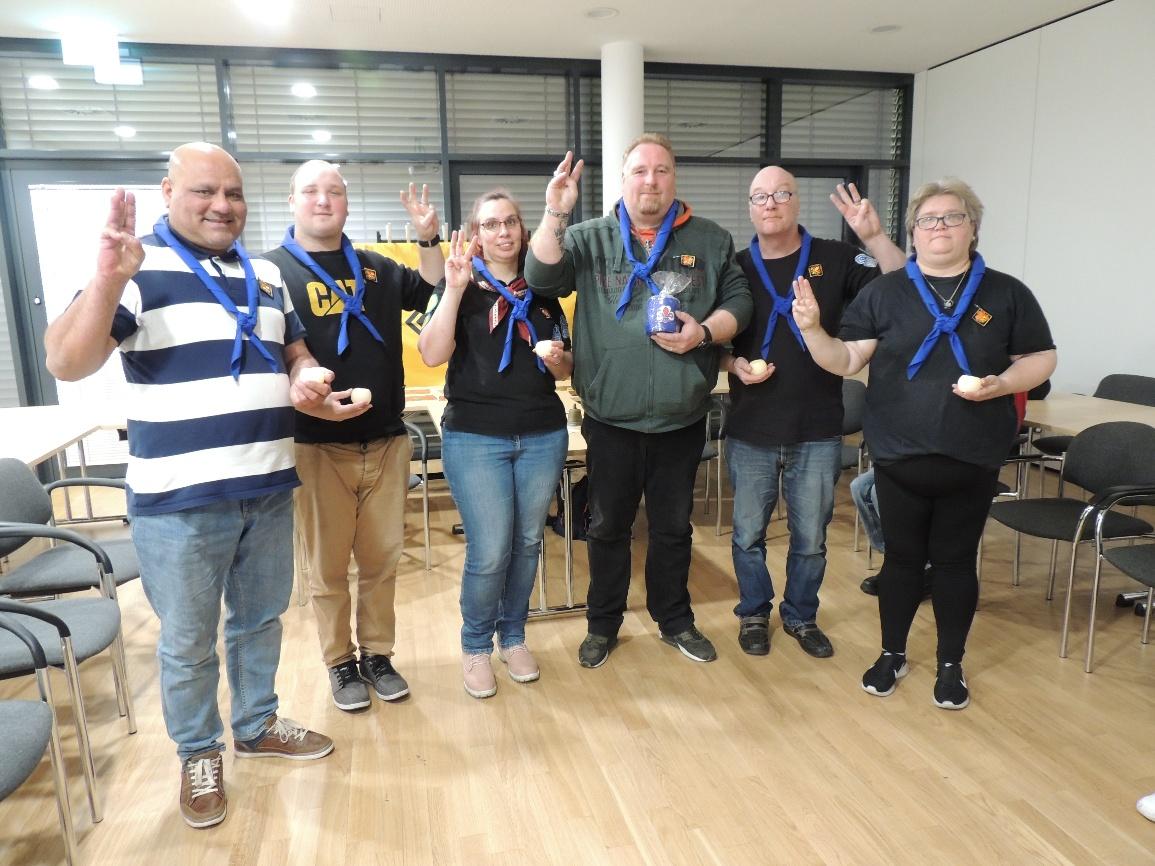 Gruppenbild: 6 Personen mit blauen Halstüchern stehen nebeneinander im Gruppenraum im Haus der Begegnung. Alle zeigen mit der Hand den Pfadfindergruß: Zeige-, Mittel- und Ringfinger werden nach oben gestreckt, während der Daumen auf den kleinen Finger gelegt wird.