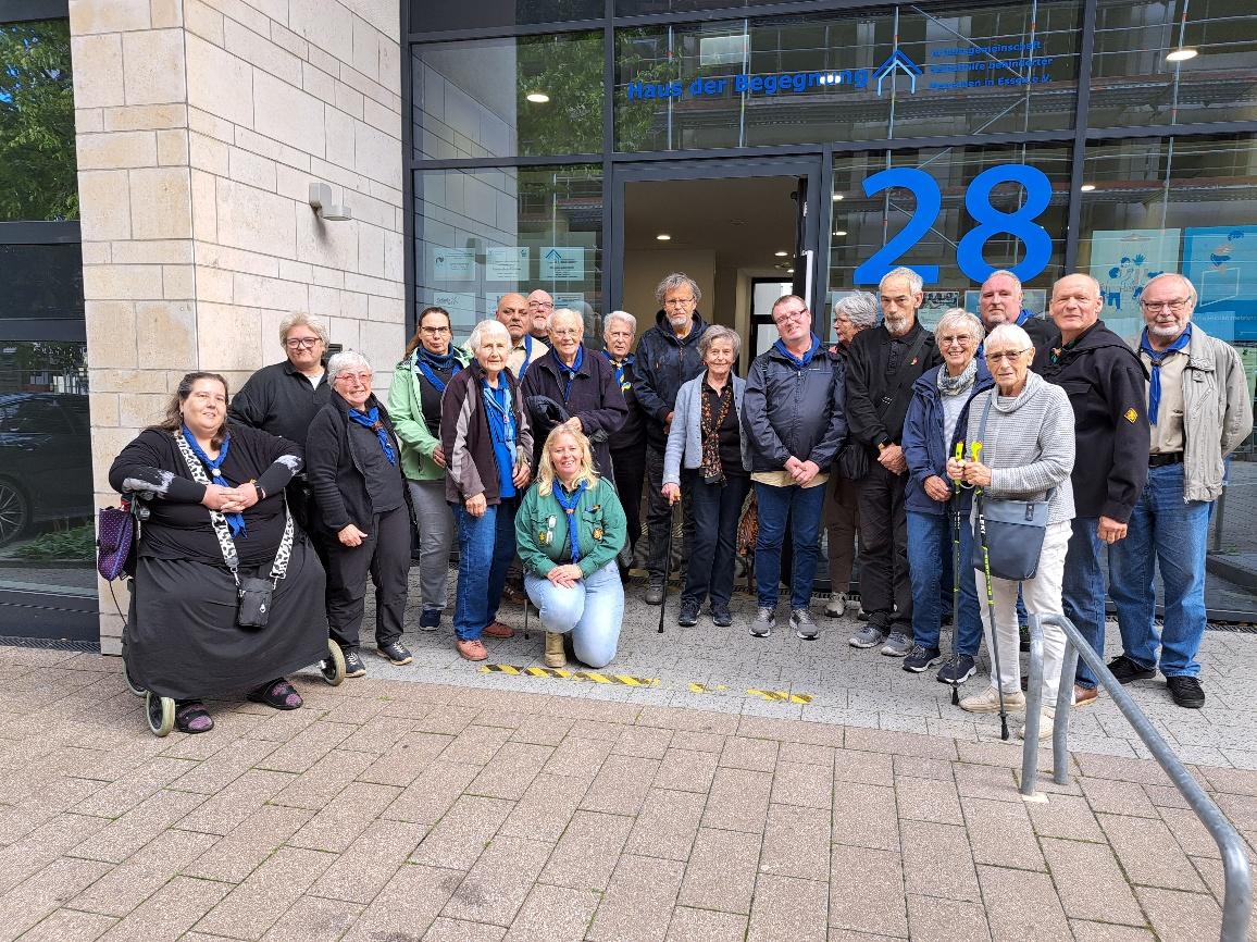 Gruppenbild vor dem Haus der Begegnung in Essen. 20 Personen sind zu sehen. Einige tragen unterschiedliche Pfadfinderbekleidung. Es gibt schwarze, grüne und beige-farbene Pfadfinderhemden. Einige tragen ein blaues Halstuch. Eine Person nutzt einen Rollator. Eine Person kniet vor der Gruppe.