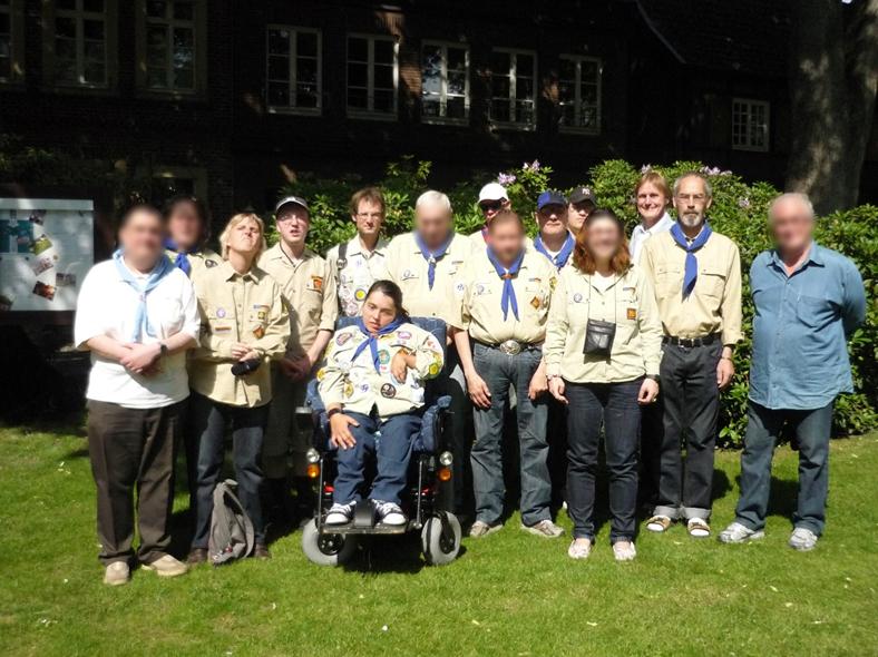 Gruppenbild von der Pfingstfahrt in 2012 nach Nottuln: 15 Pfadfinderinnen und Pfadfinder in einer beige-farbenen Pfadfinderkluft und mit einem blauen Halstuch. Die Pfadfinderkluften haben viele bunte Aufnäher. Eine der Teilnehmenden nutzt einen Rollstuhl. Im Hintergrund grüne Sträucher und ein Gebäude.