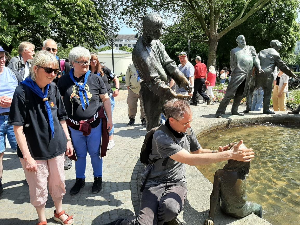 Am Brunnen „Kreislauf des Geldes“ in Aachen. 9 Personen stehen in der Nähe des Brunnens. Eine Person sitzt auf dem Brunnenrand und ertastet eine Brunnenfigur.