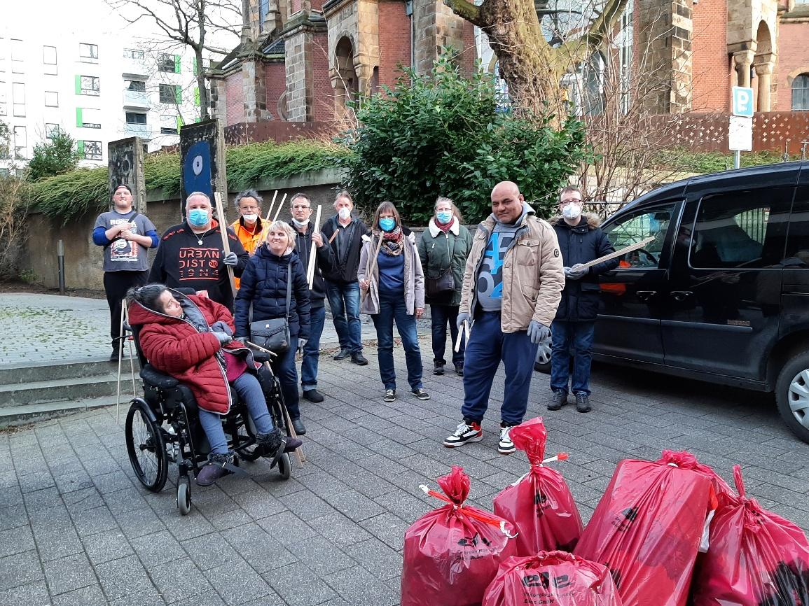 Gruppenbild: 11 Personen stehen auf dem Parkplatz des Hauses der Begegnung in Essen. Davon nutzt eine Person einen Rollstuhl. Sie halten Greifzangen aus Holz in der Hand. Im Vordergrund stehen 5 volle pinkfarbene Müllsäcke mit der Aufschrift „ebe“. Im Hintergrund sieht man die Kreuzeskirche.